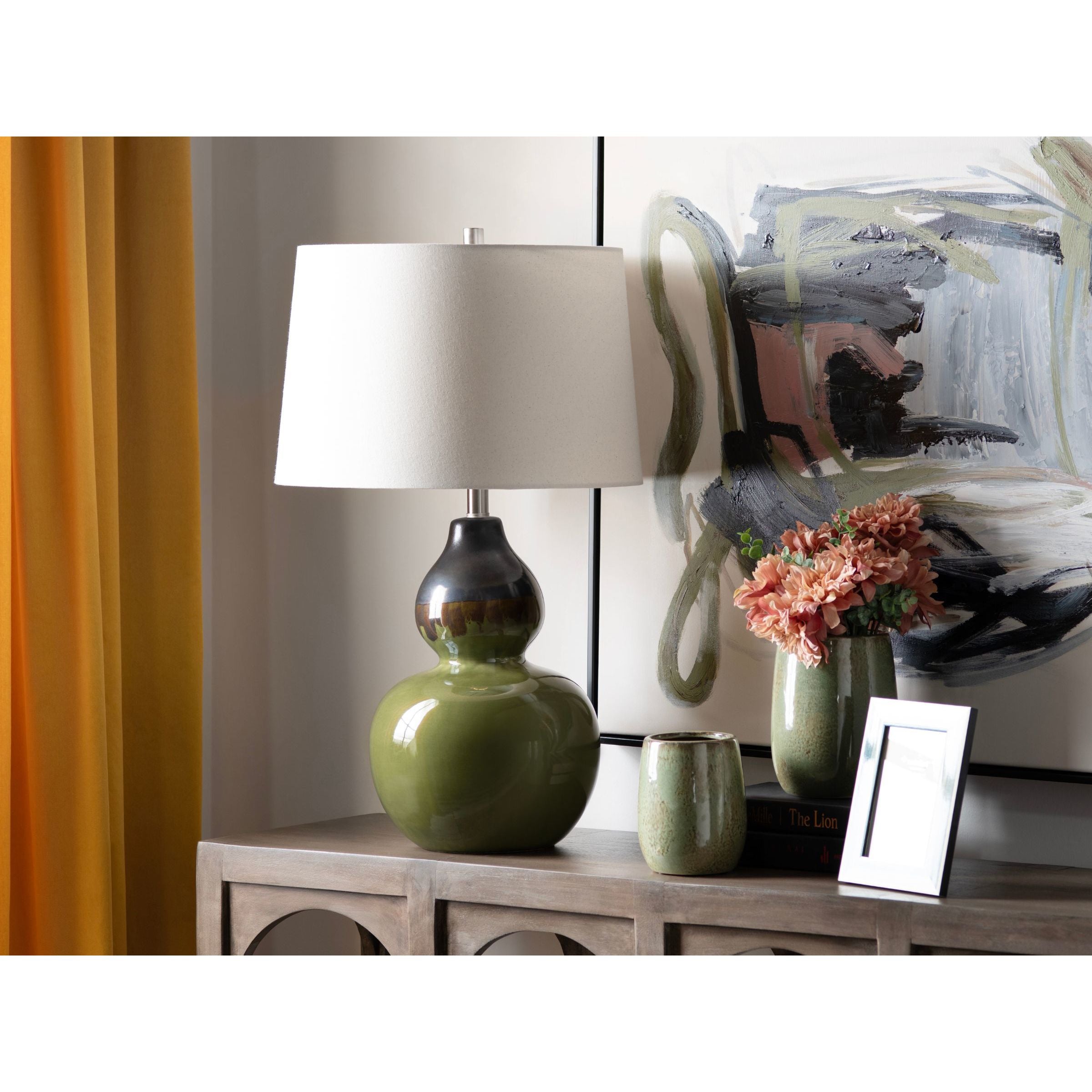 Green ceramic table lamp with white shade on console table, next to flowers and framed art.