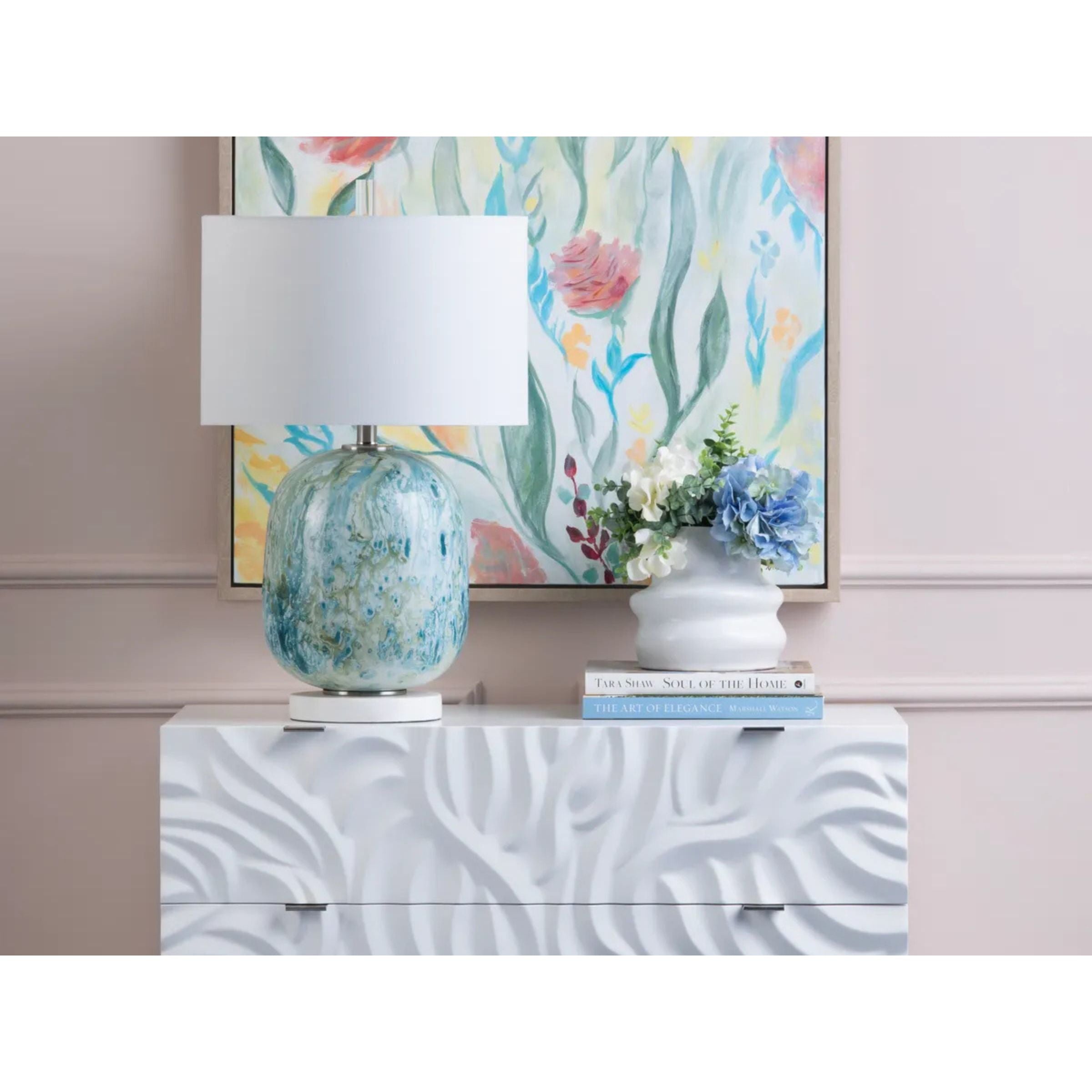 Textured white dresser with blue glass lamp, floral painting, and vase of flowers.