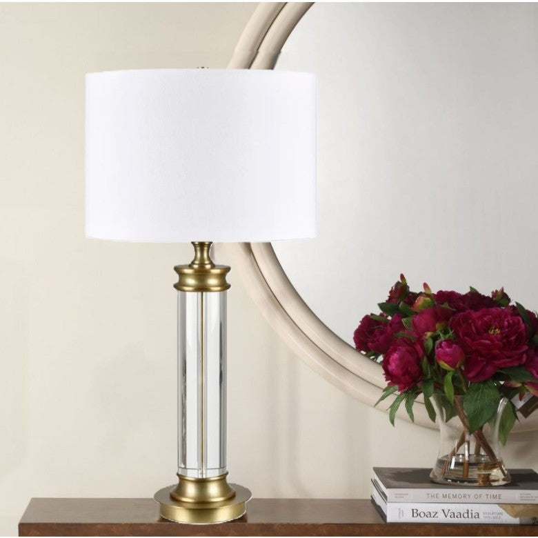 Brass and glass table lamp with white shade next to a vase of red flowers in front of a round mirror.