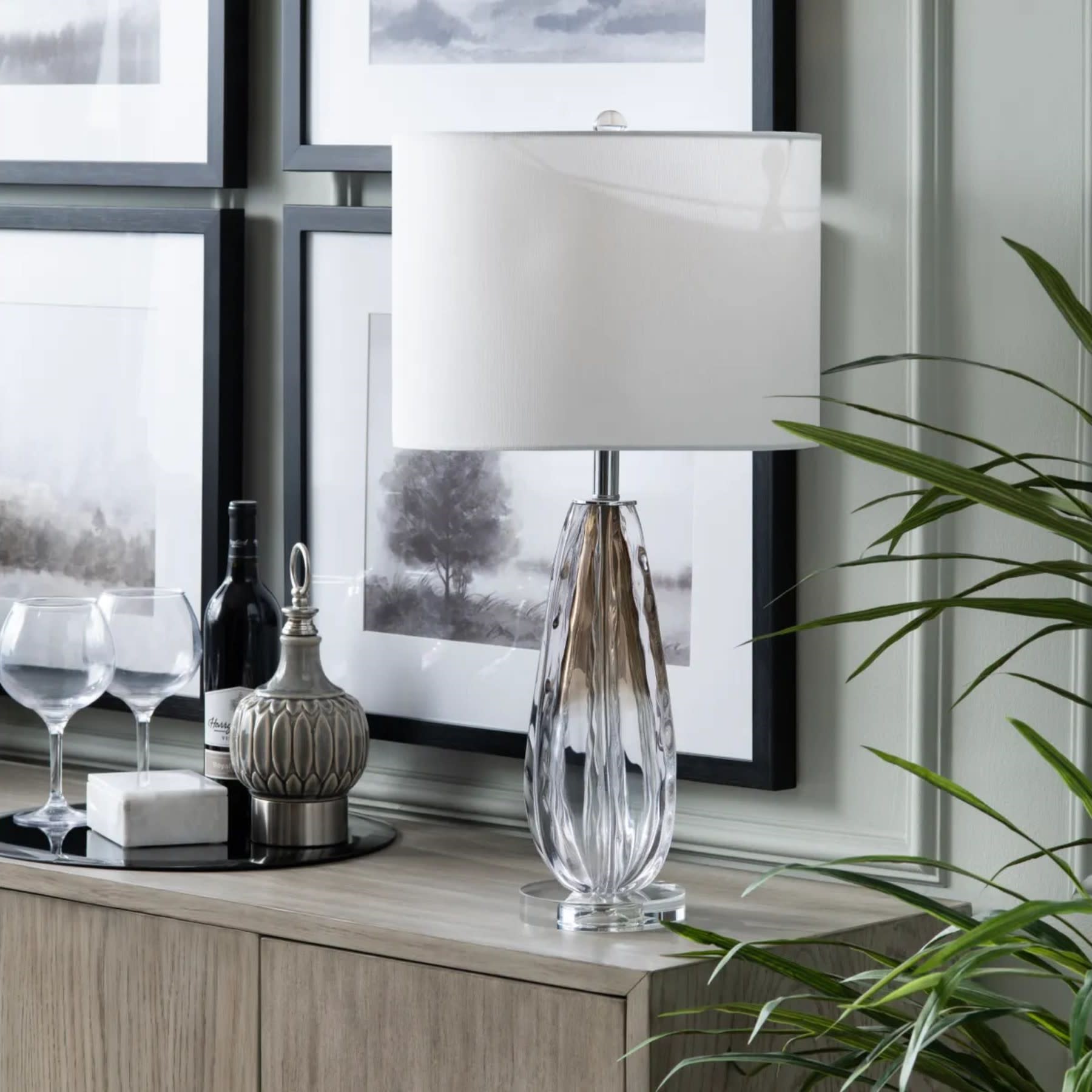 Glass table lamp with white shade on a modern wood cabinet, with decor and framed art in background.