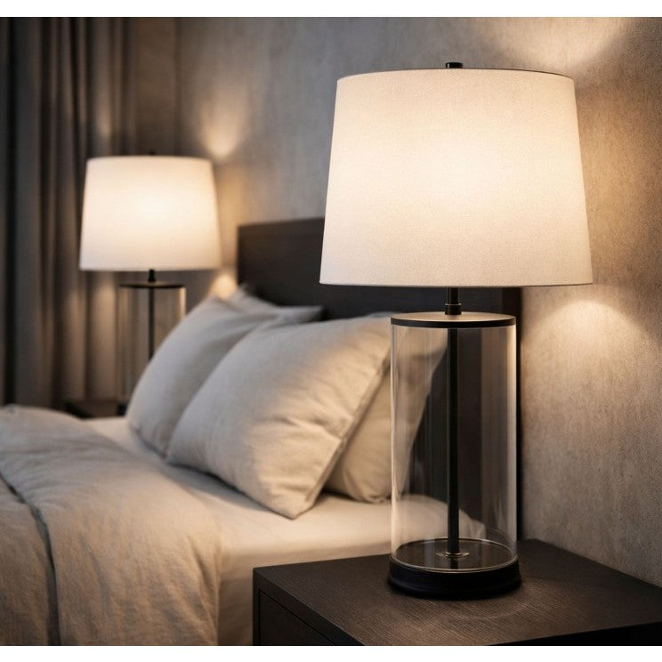 Modern bedside lamp with a cylindrical glass base and white shade on a nightstand.