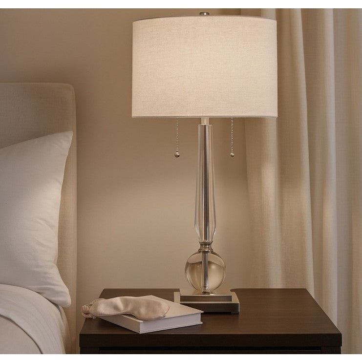 Glass table lamp with white shade on bedside table next to a bed and curtain.