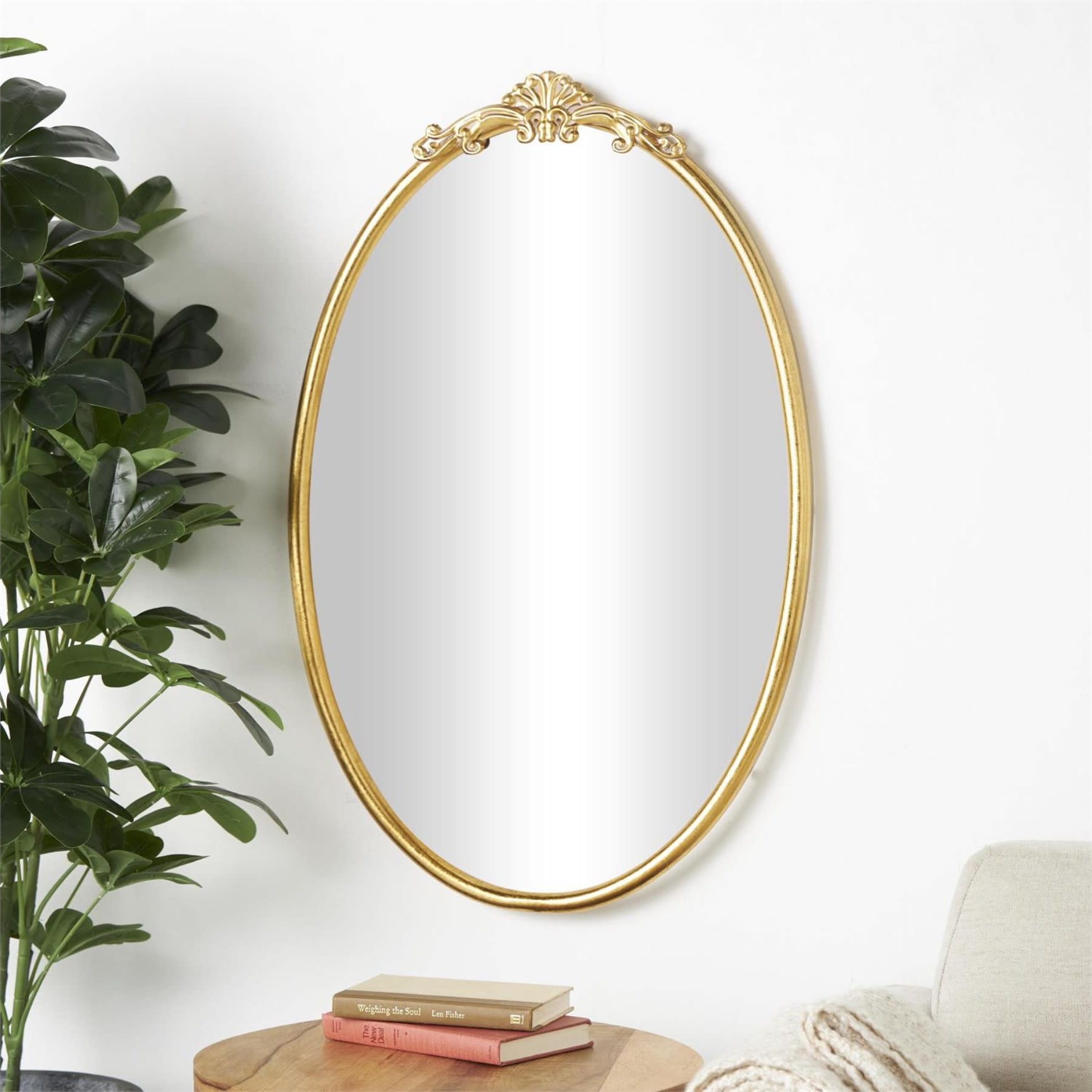 Oval gold-framed wall mirror above a small table with books and a plant.