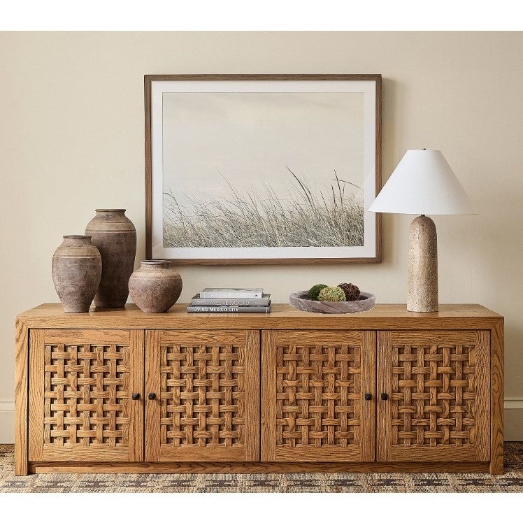 Wood sideboard with woven doors, decorated with vases, books, a bowl, and a lamp under framed art.