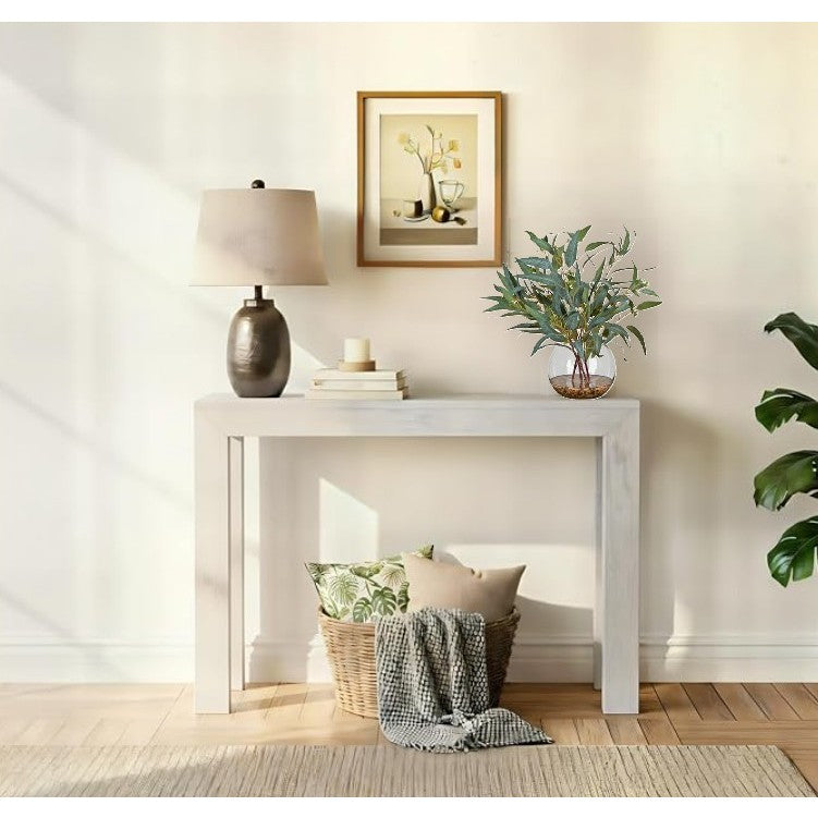 Modern console table with a lamp, framed art, plant, and basket with pillows in a bright room.