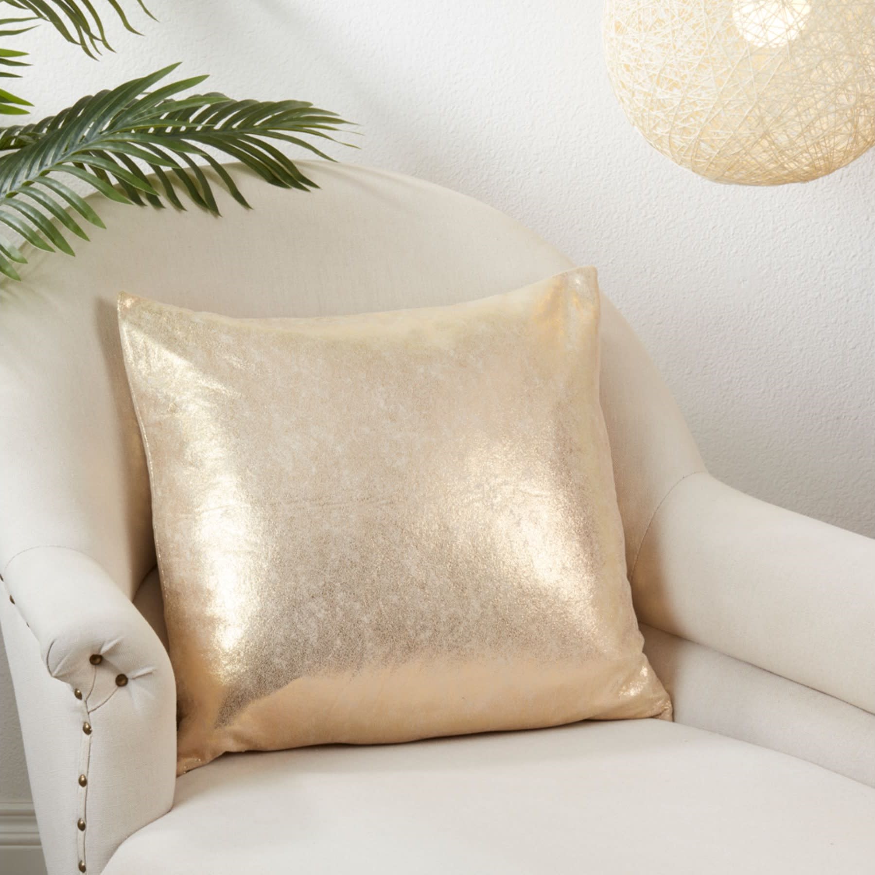 Gold metallic throw pillow on a cream armchair, with decorative plant and hanging light.
