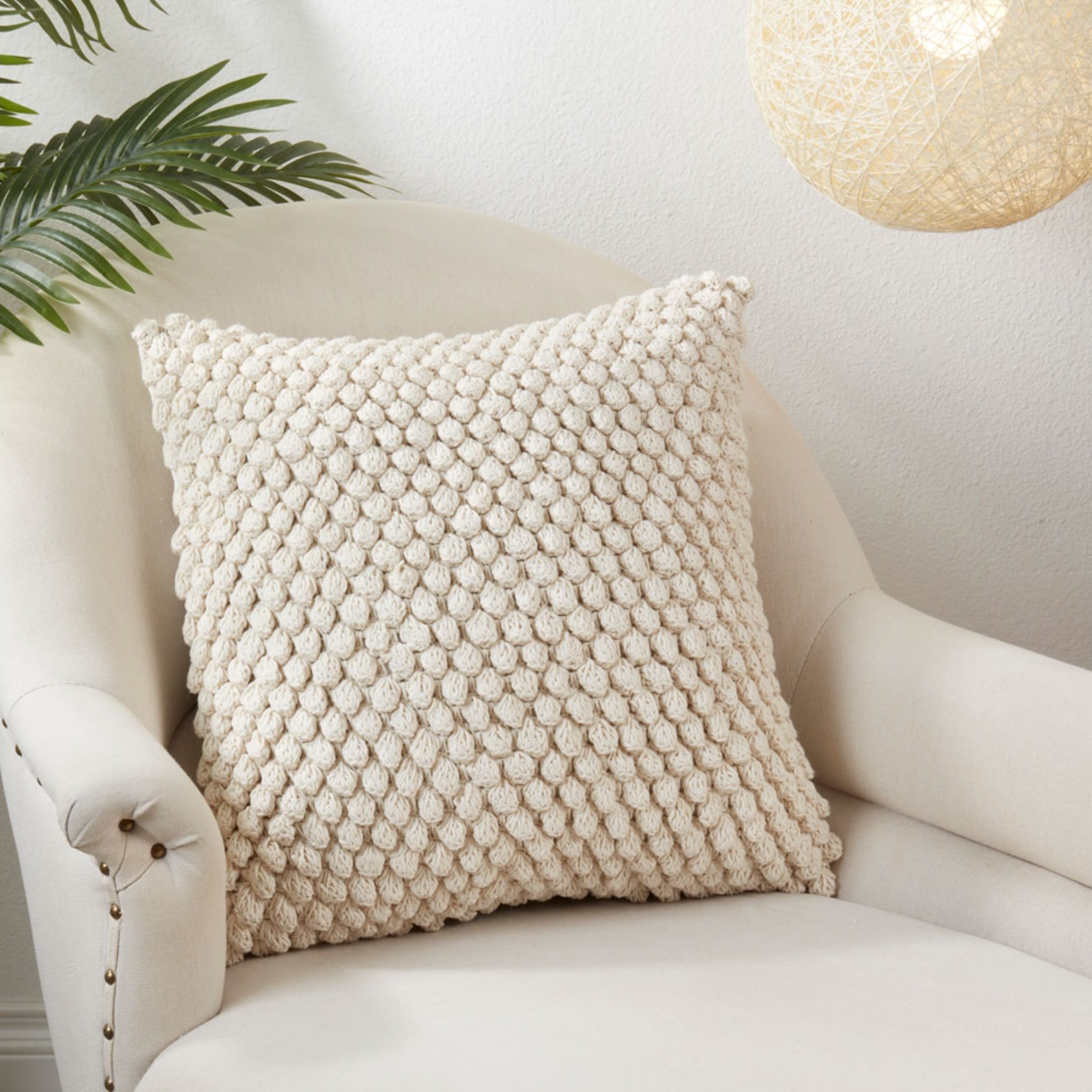 Textured cream pillow on a beige armchair, with plant and light fixture in background.