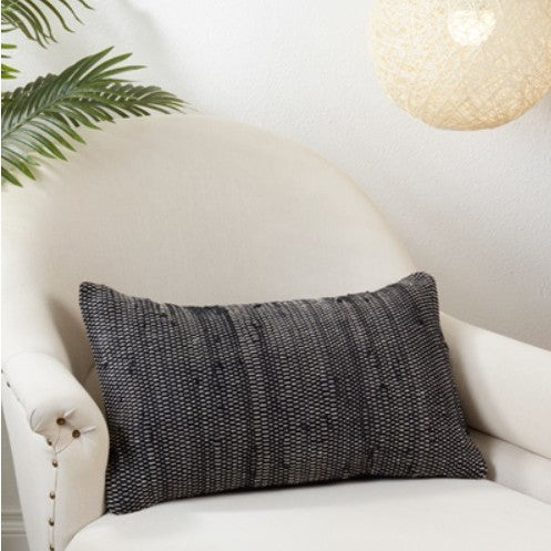 Black textured pillow on white lounge chair with a leafy plant and hanging lamp nearby.