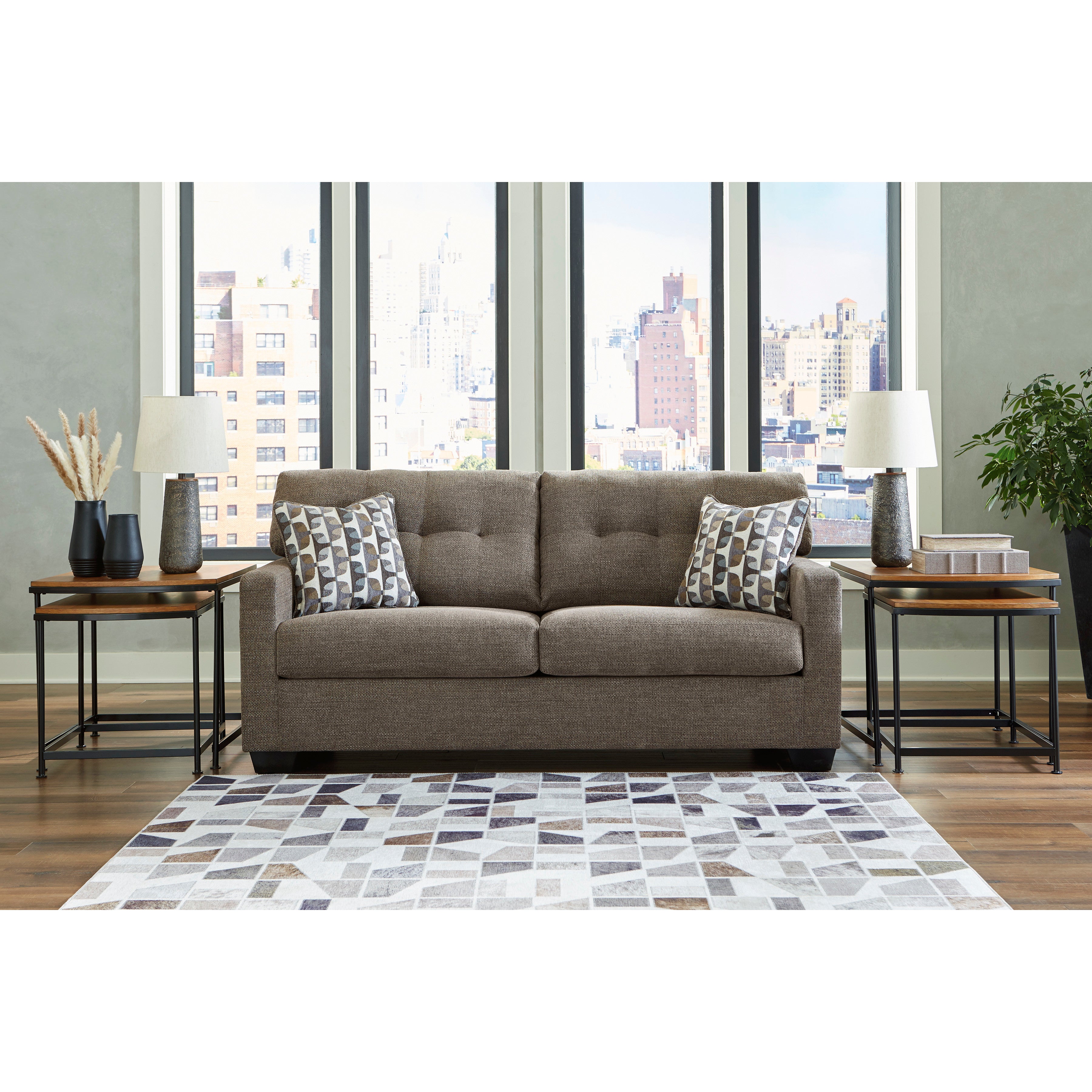 Contemporary brown sofa with two patterned pillows, side tables, and lamps in a bright living room.