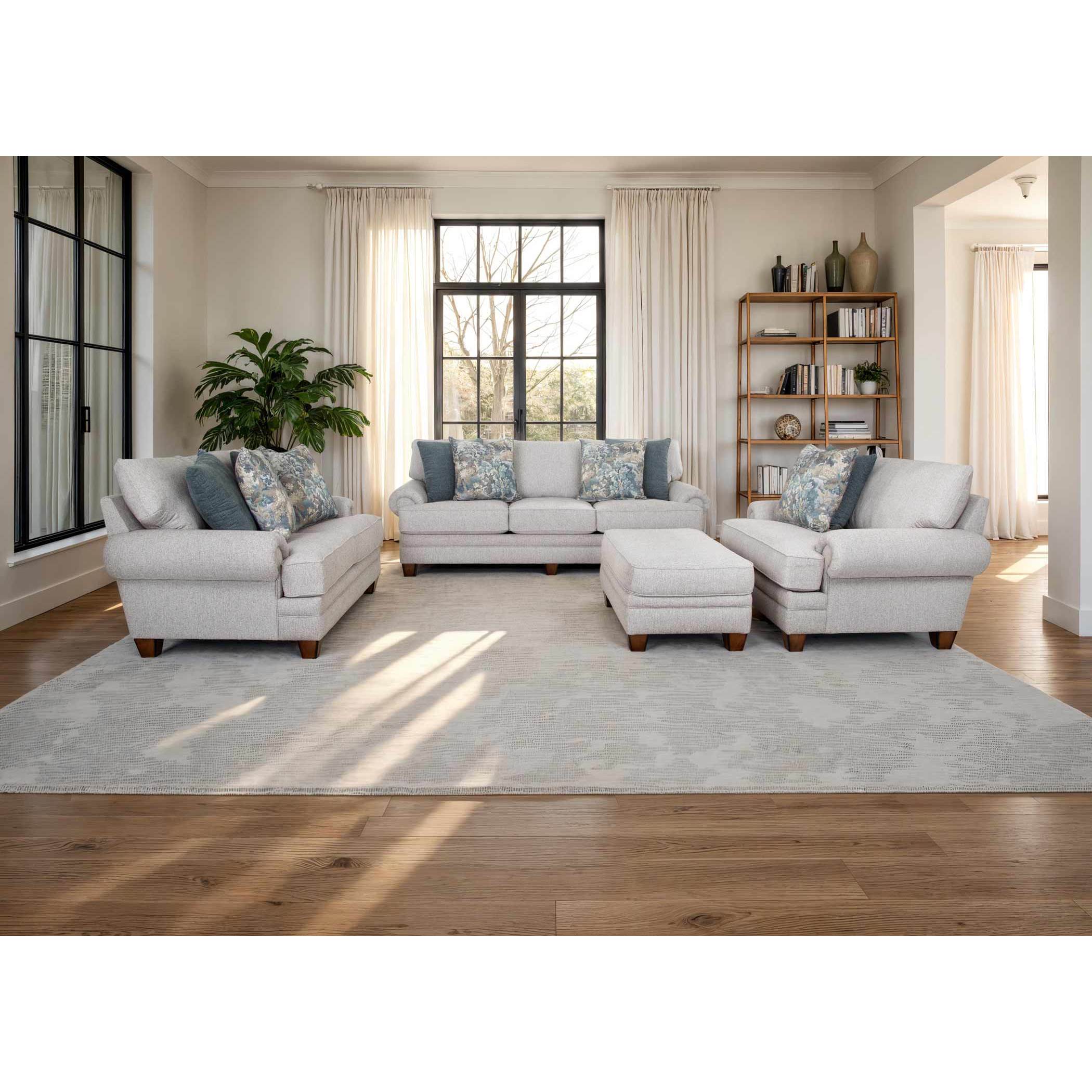 Modern living room with light sofas, accent pillows, ottoman, rug, and bookshelf near large windows.