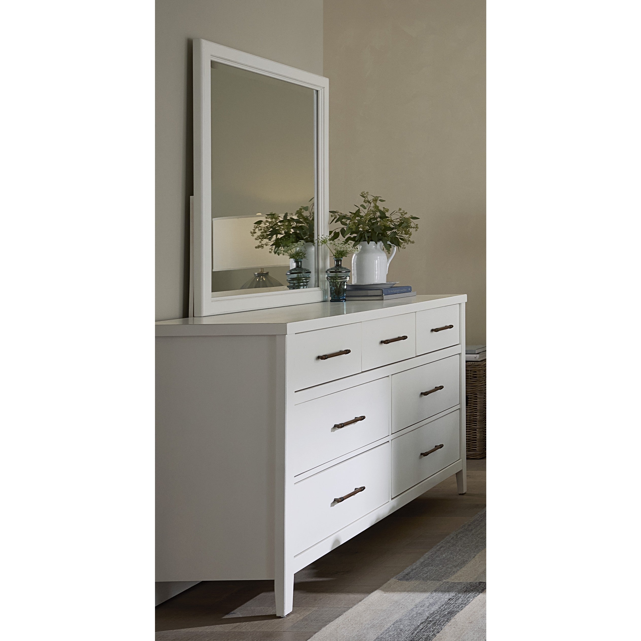 White dresser with mirror, decorated with books, plants, and vases in a bedroom corner.