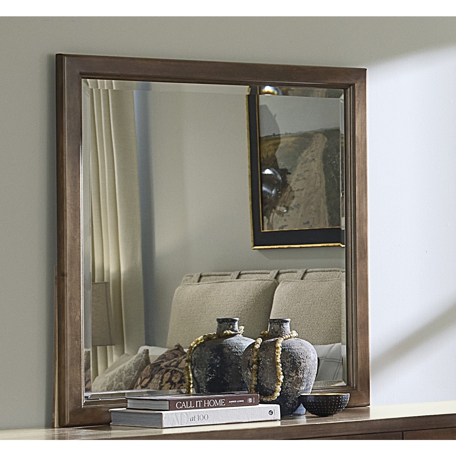 Square wall mirror with wooden frame above dresser with vases and books.