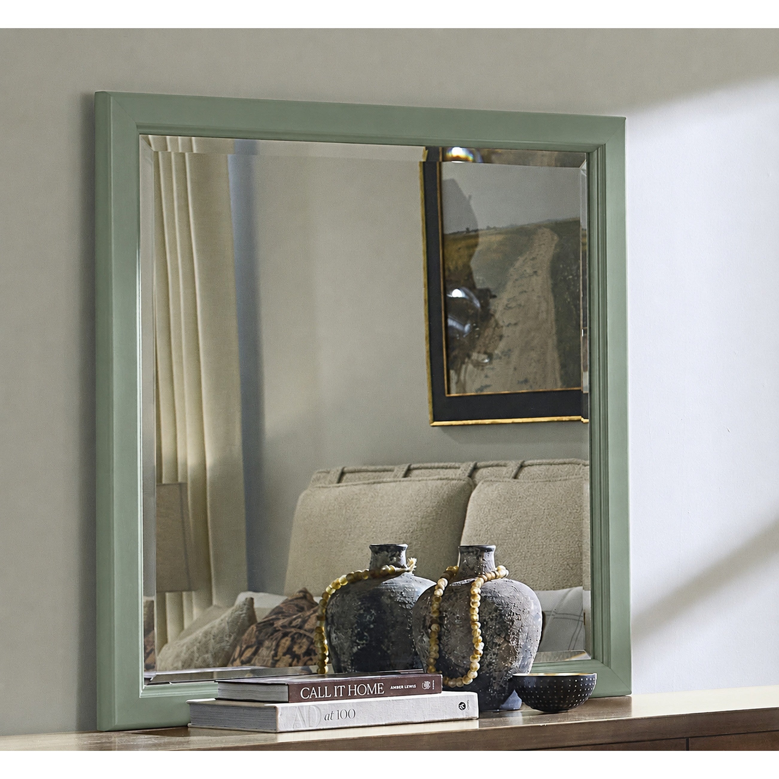 Square wall mirror with a light green frame above a console table with books and vases.
