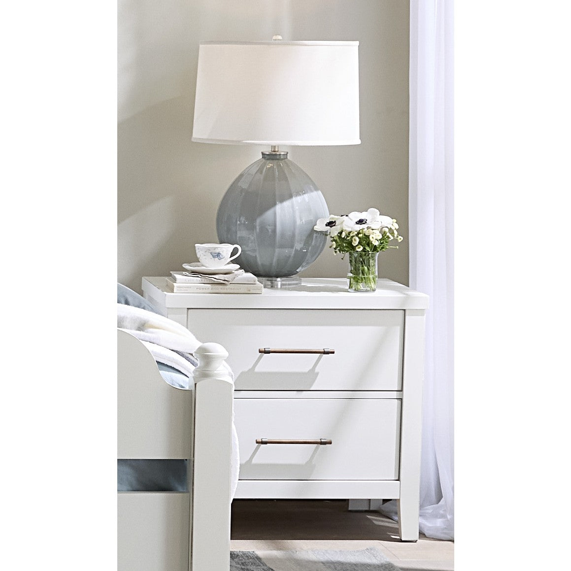White nightstand with two drawers, a lamp, flowers, and a teacup on top.
