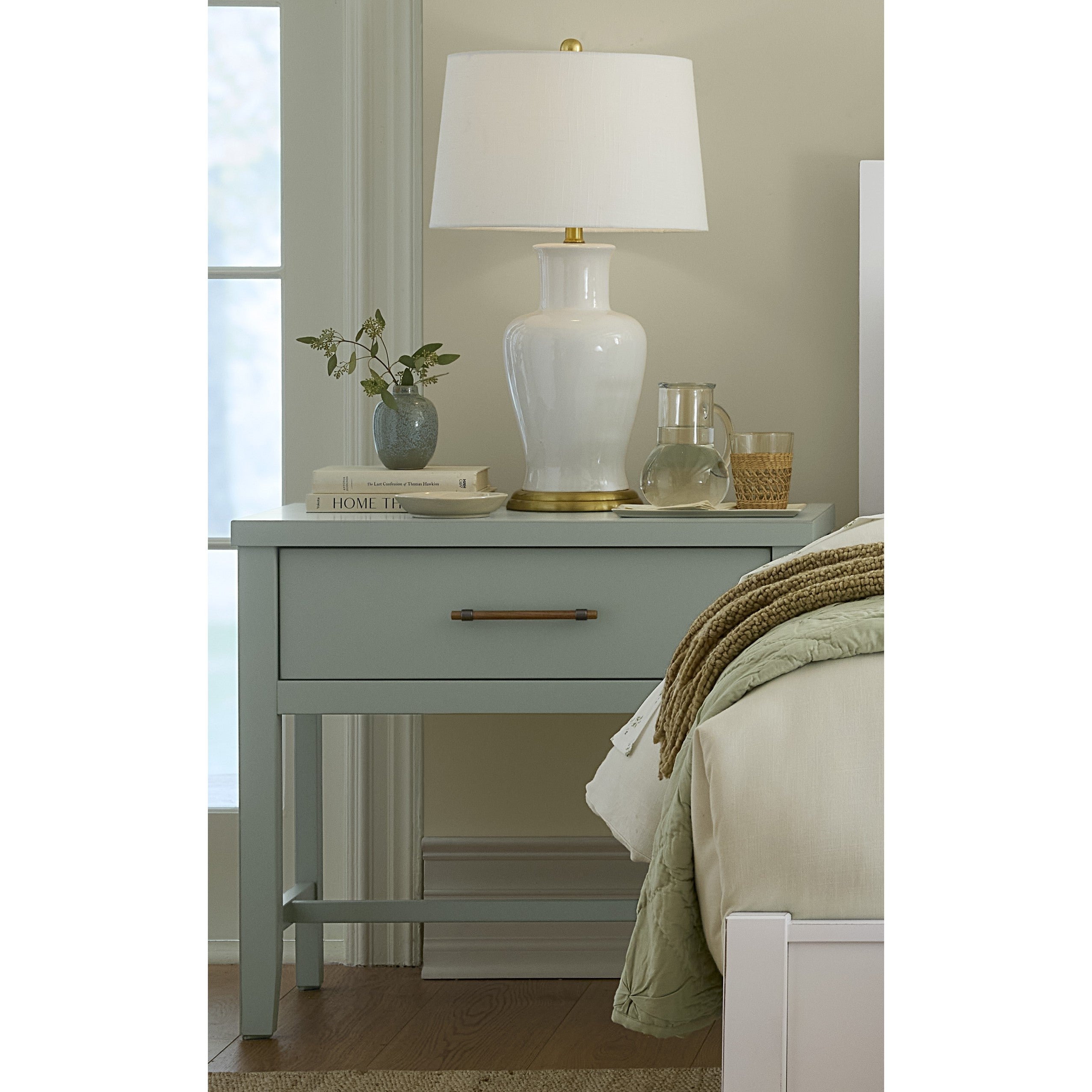 Nightstand with lamp, books, plant, glass pitcher, and glass beside a bed.