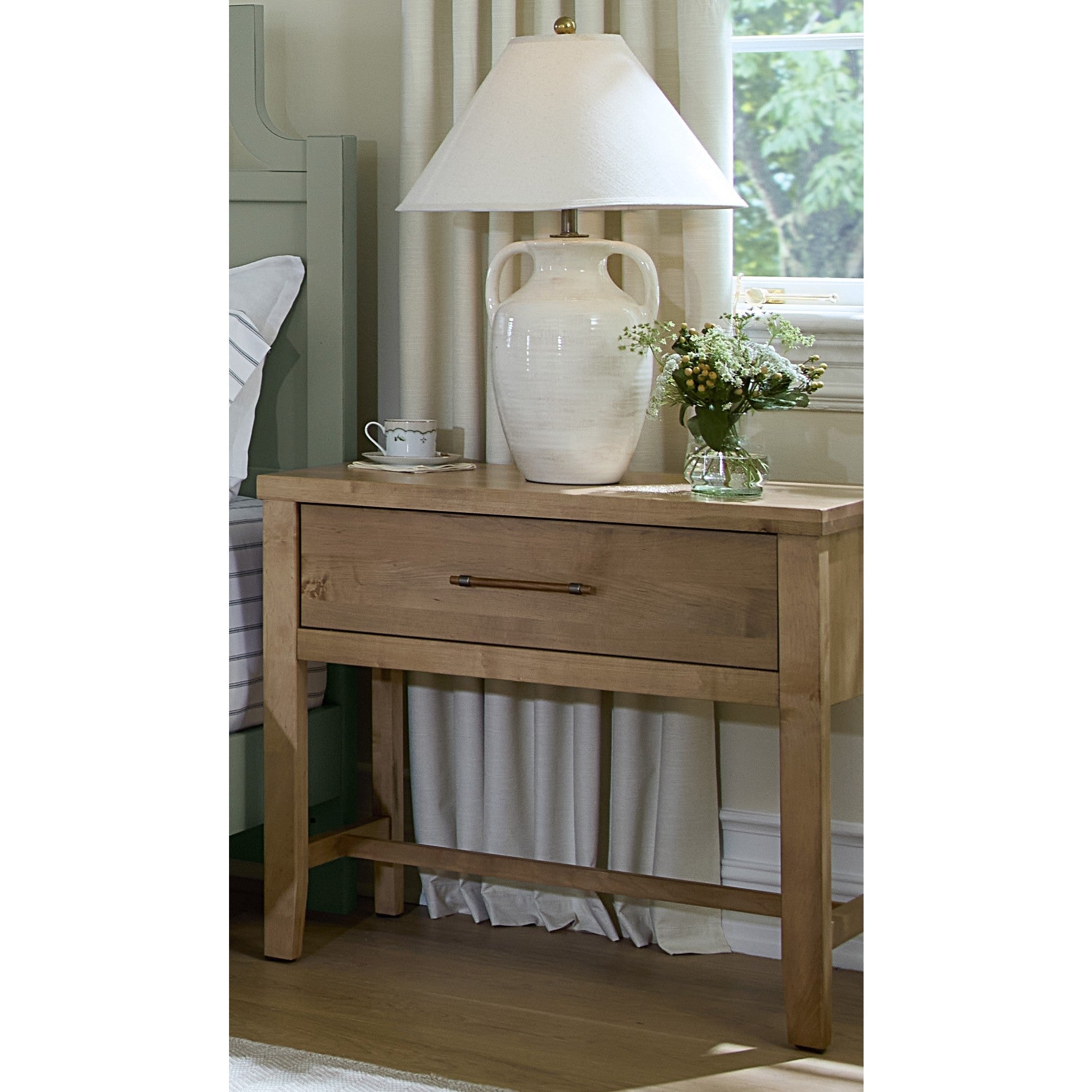 Wood nightstand with drawer, lamp, cup, and flowers in a bedroom setting.