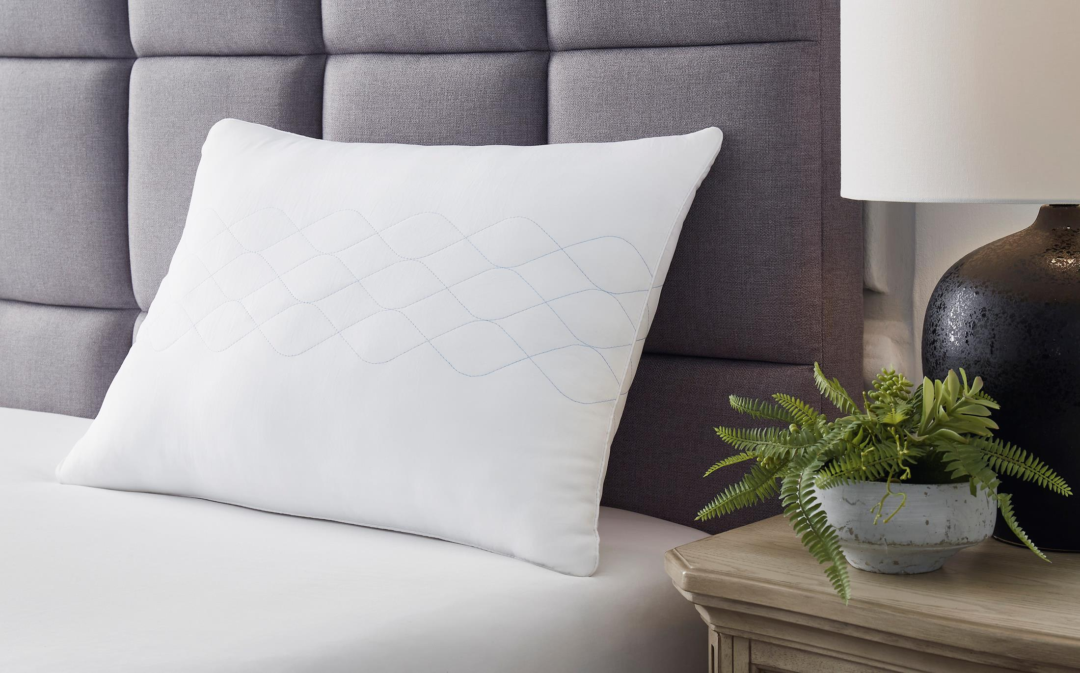 White pillow on a bed next to a gray headboard and a small plant on a nightstand.