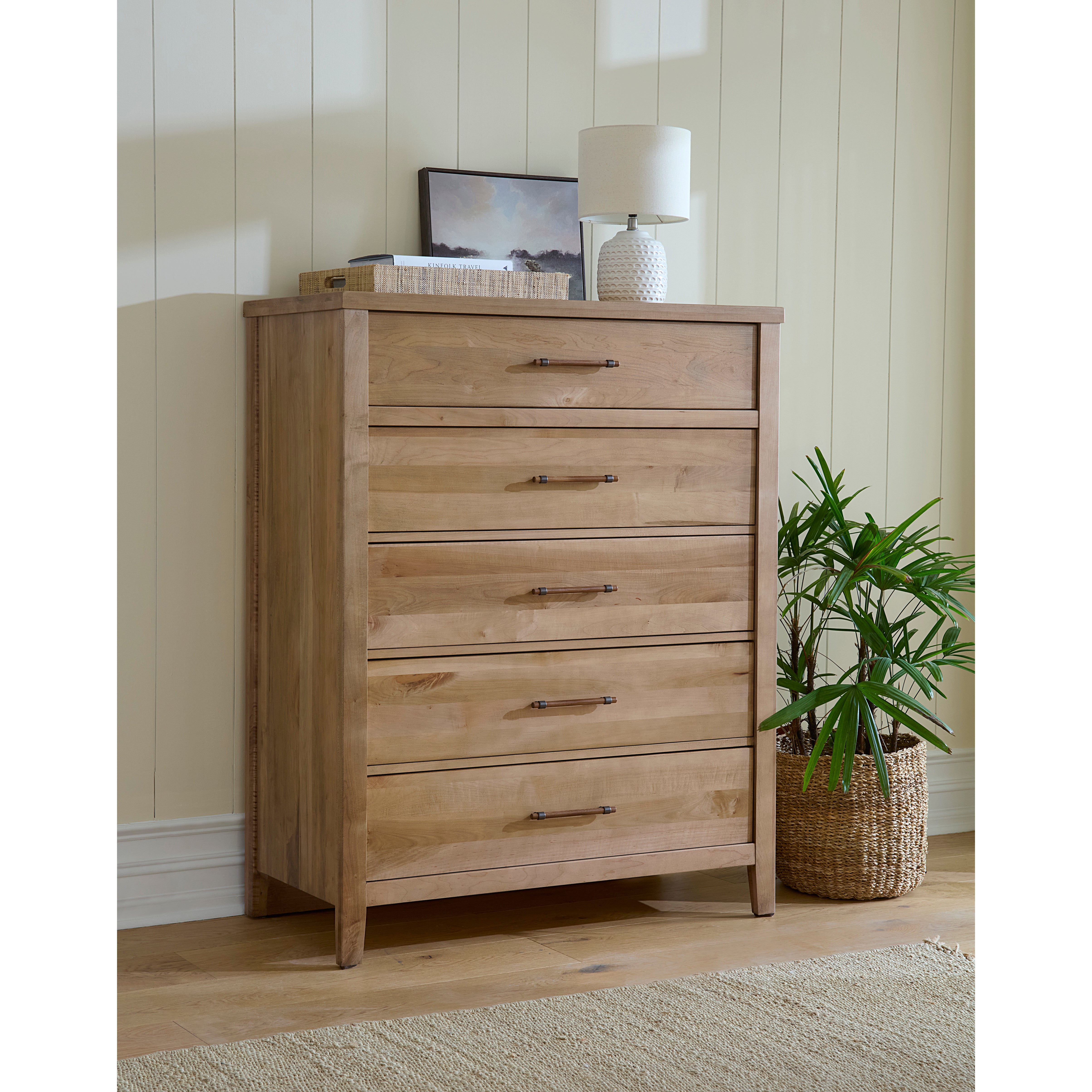 Natural wood dresser with five drawers, lamp, framed art, and a plant on the side.
