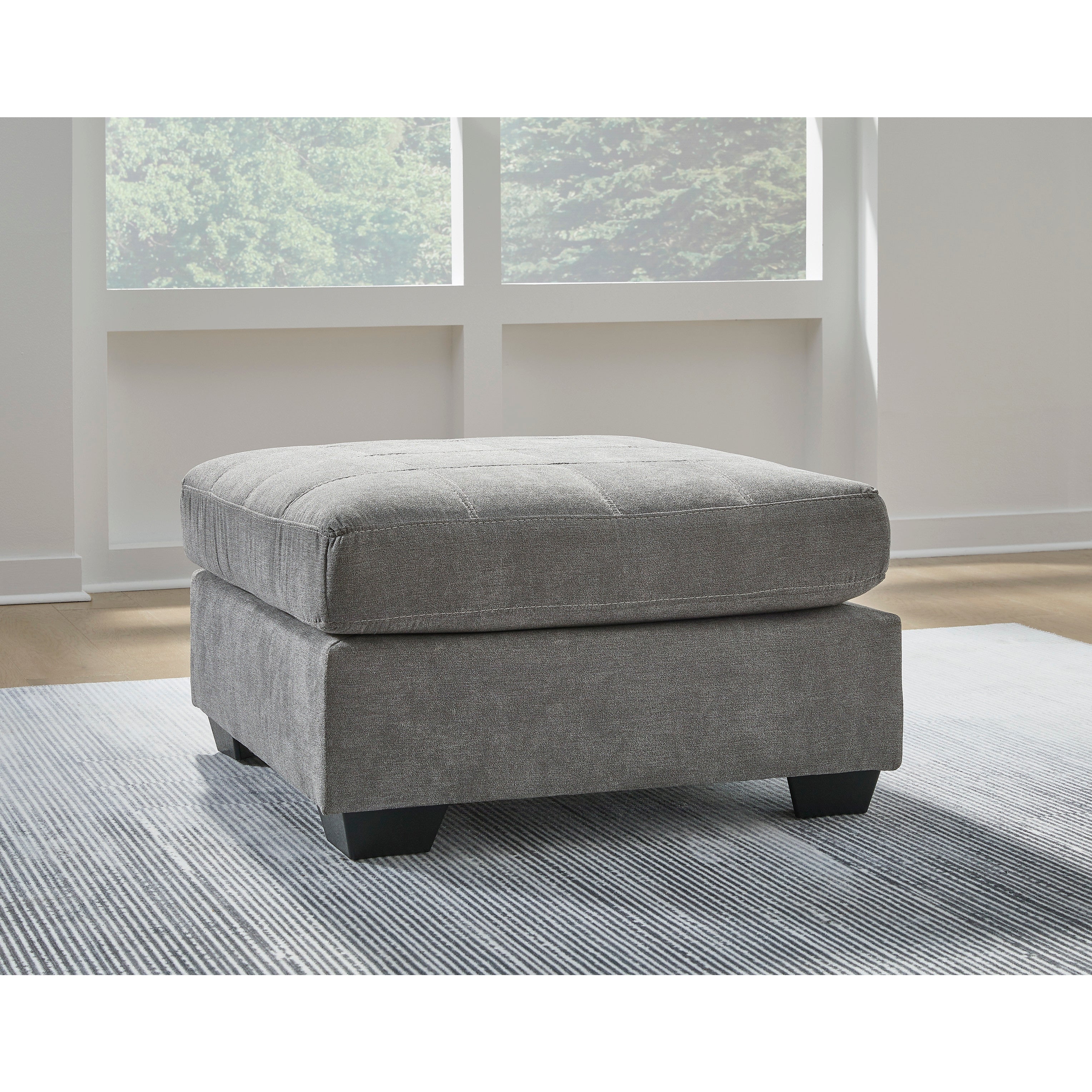 Gray upholstered ottoman with short legs on a striped rug in front of bright windows.