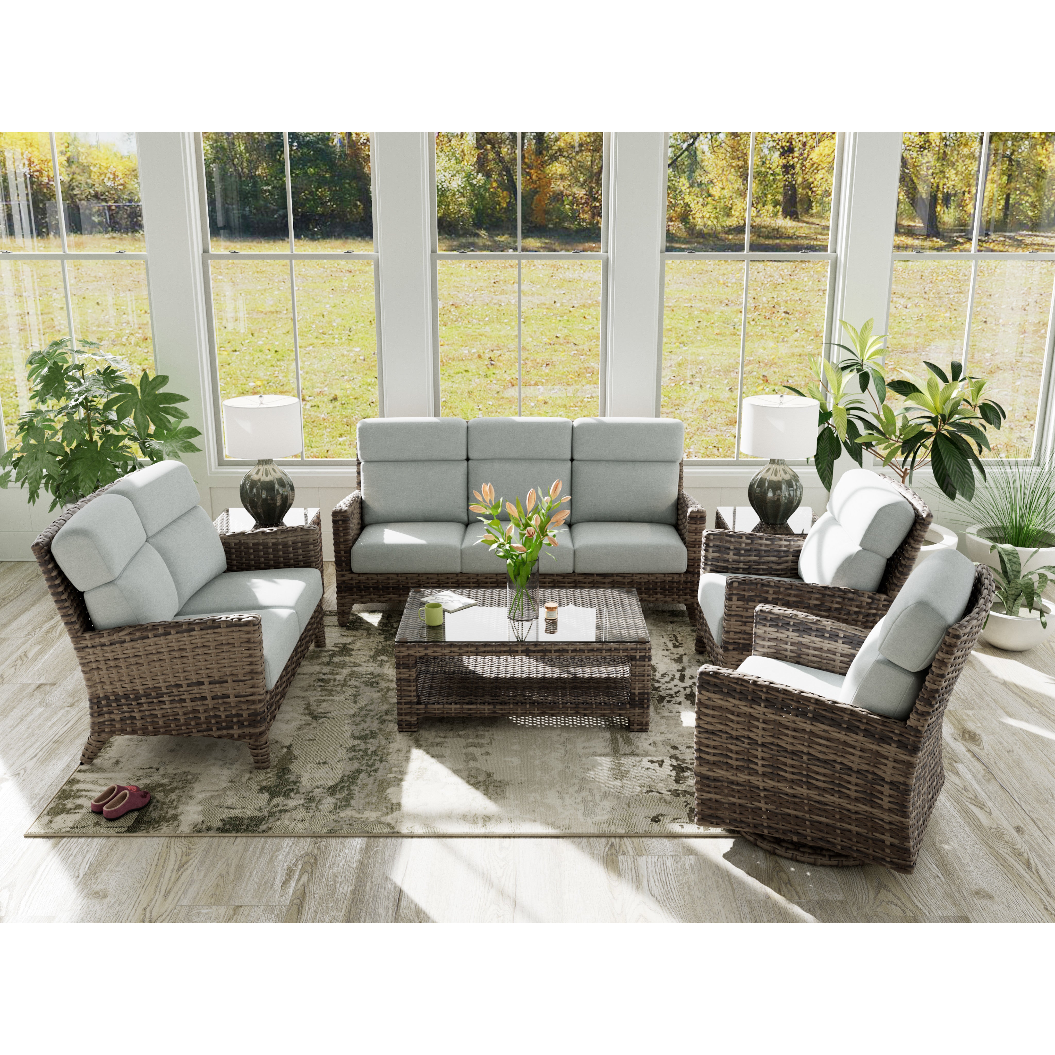 Wicker patio furniture set with light gray cushions in a sunroom with large windows and green plants.