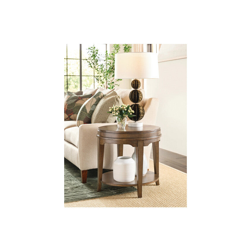 Round wood end table with lamp and vase beside a beige sofa in a living room.