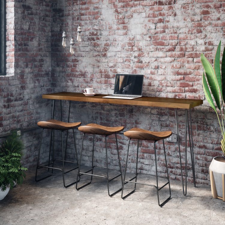 Modern bar table with three stools, laptop, and decor against an exposed brick wall.