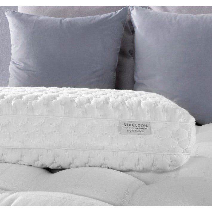 White textured pillow labeled 'Aireloom Nimbus Visco' on a bed with two gray pillows.